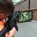 A child in an orange shirt, immersed in a laser tag game, aims a black toy gun while standing indoors near a graffitied dumpster, with the buzz of a birthday party and the motion of peers partially visible in the background.