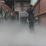 Two people face each other down a foggy, graffiti-lined hallway in an industrial setting; one points a camera while the other, ready for a laser tag corporate team building activity, stands near a wall.