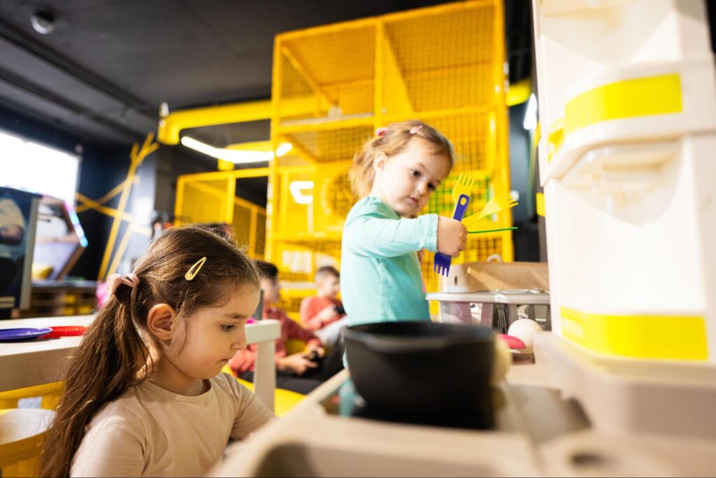 Two young girls play with toy kitchen equipment in a brightly lit indoor play area in Florham Park, NJ, perfect for fun indoor summer activities, with cheerful yellow play structures in the background.