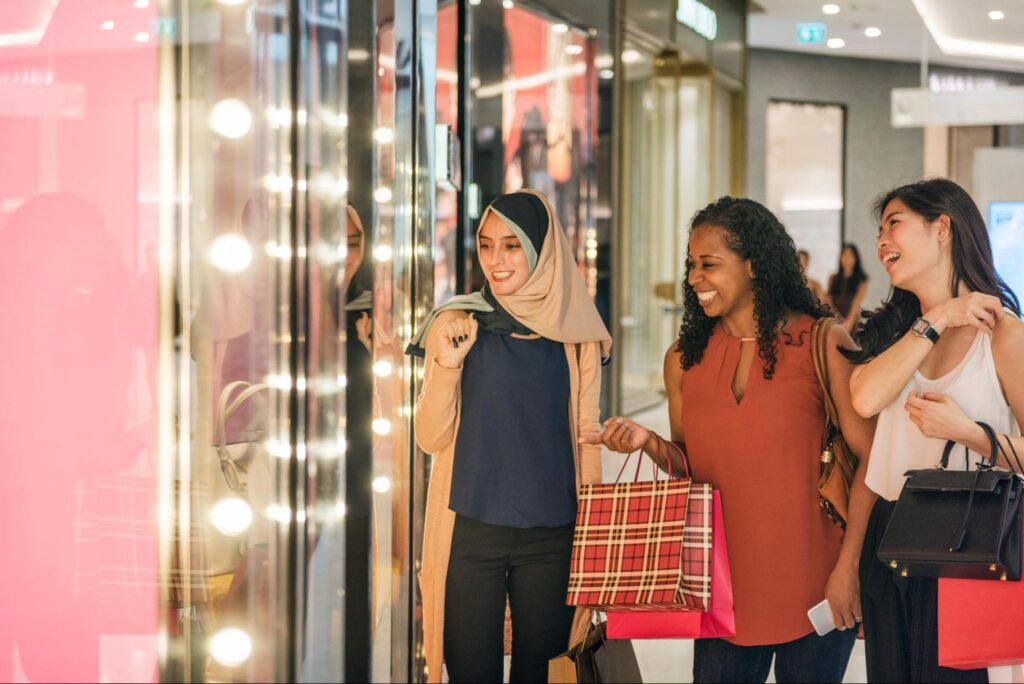Three women are standing in a shopping mall in Princeton, smiling and looking at a brightly lit store window while holding shopping bags—enjoying one of the best summer things to do on their adventure list.