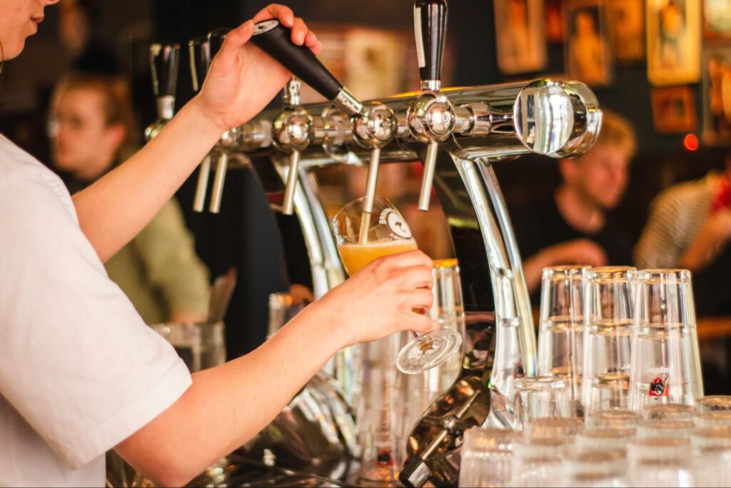 A person fills a glass with draft beer from a tap in a bar, with stacked glasses and patrons visible in the background—perfect for unwinding after Princeton summer activities or as part of your own Princeton adventure.
