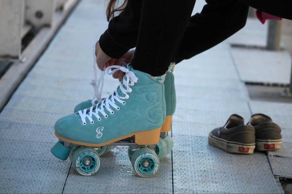 A person ties the laces of teal roller skates while wearing black pants; a pair of slip-on sneakers sits nearby, perfect for indoor summer activities in Florham Park NJ.
