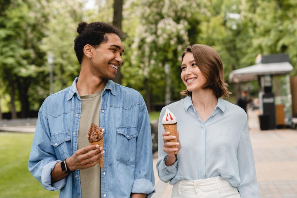 Two people walk outdoors in a park, smiling at each other while holding ice cream cones. Trees and a food kiosk are visible in the background, making it one of the fun date ideas for those seeking unique New Jersey activities.