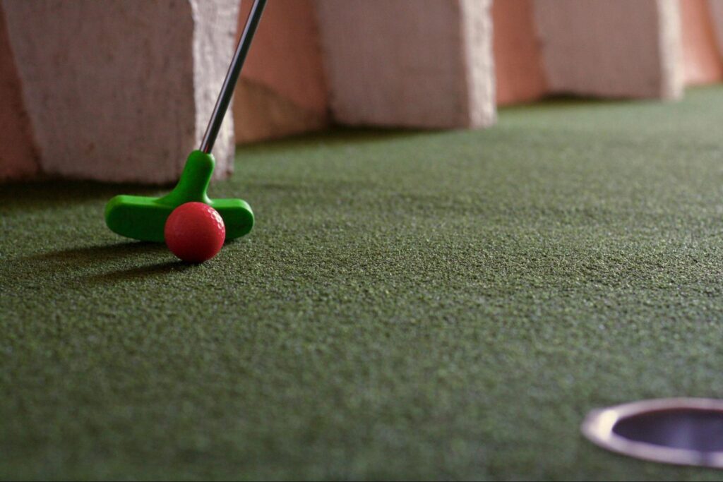 A green mini-golf putter is positioned behind a red golf ball on artificial turf, aiming toward a cup—perfect for fun activities or double date ideas in New Jersey.