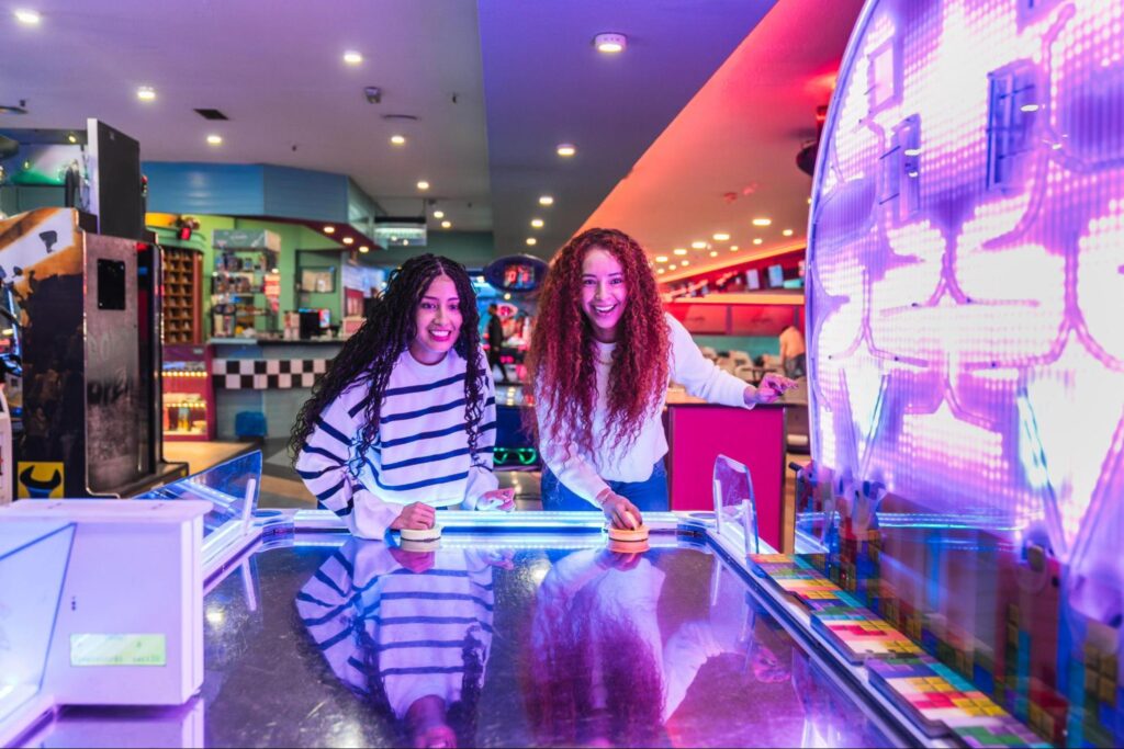 Two young women enjoying air hockey in an arcade, smiling and focused on the game amidst colorful lights—perfect for anyone seeking double date ideas or fun activities in New Jersey.