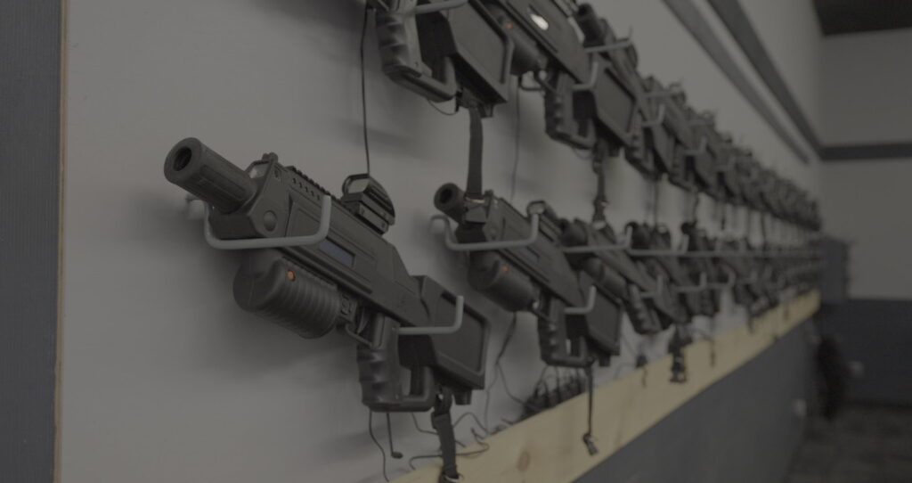 A row of black replica guns mounted on a wall, each secured with brackets and connected by wires, creates an intriguing setting for indoor summer activities in Fairfield NJ—perfect to beat the heat in a dimly lit room.