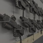 A row of black gaming rifles is mounted on a wall, secured with brackets and cables, in a dimly lit room—perfect for those seeking fun indoor summer activities.