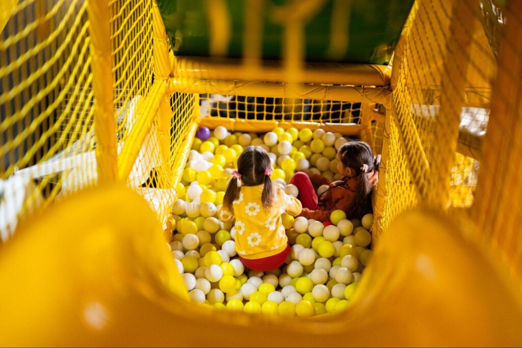 Two young children sit in a ball pit filled with yellow and white balls, surrounded by yellow netting in an indoor play area—one of the best Indoor Summer Activities Fairfield NJ has to offer!.
