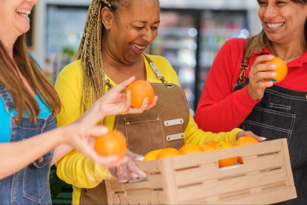 Three women wearing aprons stand together, smiling and holding oranges from a wooden crate in a store—a fresh team activity that would inspire unique corporate event ideas for Tac Ops NJ.