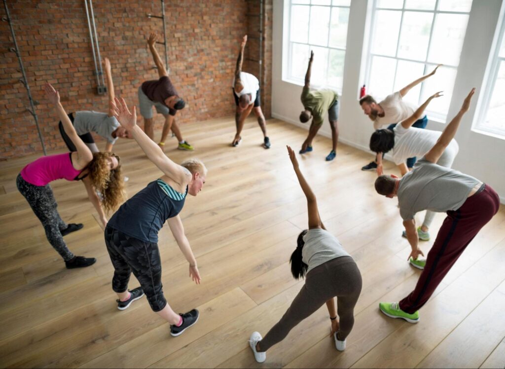 A group of people in athletic wear are standing in a studio, stretching with arms extended and leaning to the side—a great example of corporate event ideas offered by Tac Ops NJ.