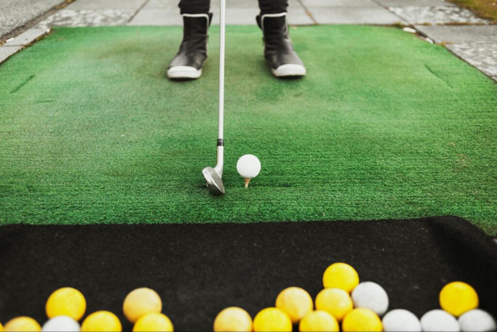 A person stands on artificial turf in Fairfield NJ, preparing to hit a white golf ball on a tee with a golf club—one of the best indoor summer activities to beat the heat, with several yellow and white balls nearby.
