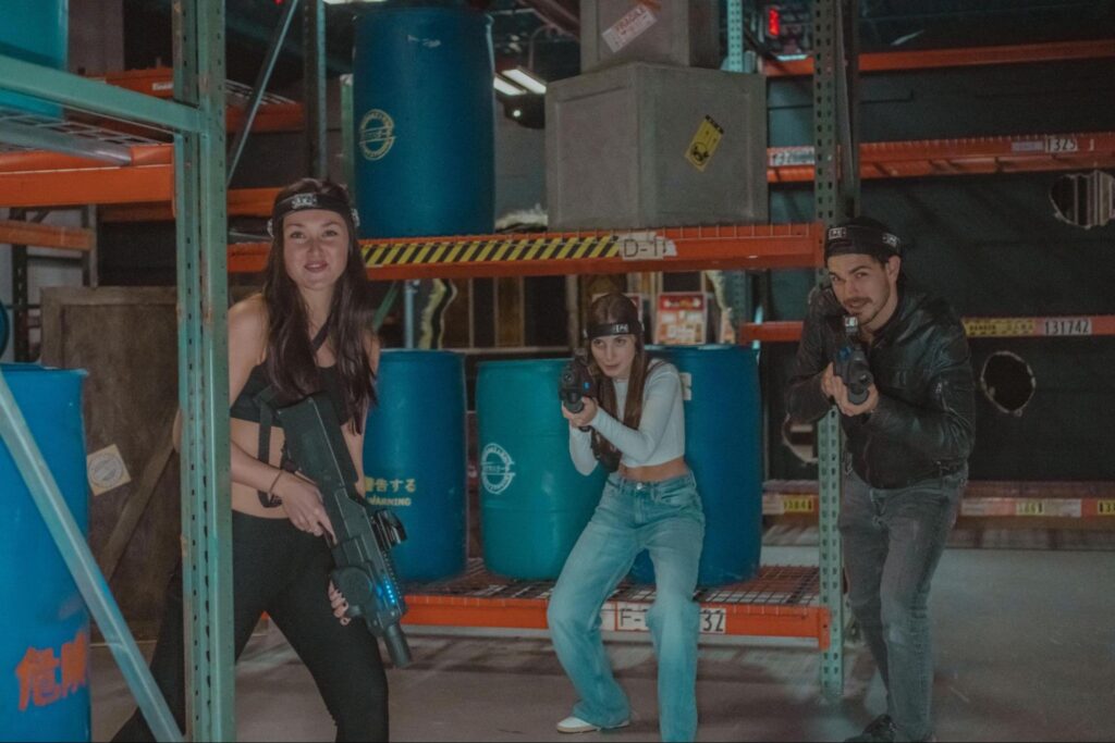 Three people in casual clothing, holding laser tag guns, pose in a warehouse-style arena with blue barrels and metal shelves at Tac Ops NJ—perfect for team building or unique corporate event ideas.