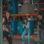 Three people in casual clothing, holding laser tag guns, pose in a warehouse-style arena with blue barrels and metal shelves at Tac Ops NJ—perfect for team building or unique corporate event ideas.