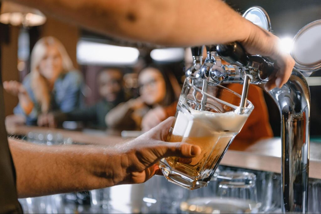 A bartender pours a glass of draft beer from a tap at one of the top NJ locations, with people sitting and talking in the background—perfect for those seeking unique corporate event ideas.