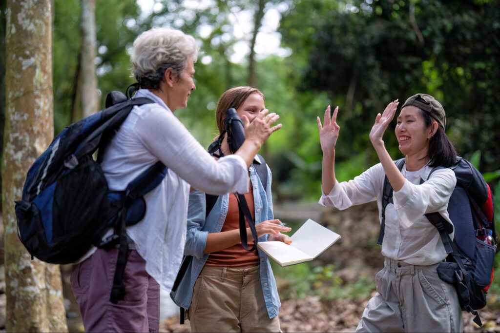 Three people with backpacks stand in a forest, smiling and high-fiving while one holds a notebook—an adventurous take on corporate event ideas inspired by Tac Ops NJ.