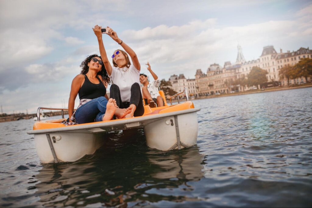 Three people enjoy a pedal boat ride on the water in New Jersey, with two taking a selfie. City buildings and trees line the background under a partly cloudy sky—perfect for those seeking fun activities or double date ideas.