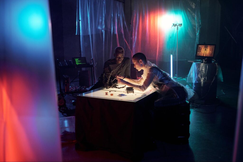 Two people work on electronic components at a lit table in a dimly lit room with colorful lights and computer screens—an ideal spot for indoor summer activities in Fairfield NJ to beat the heat.
