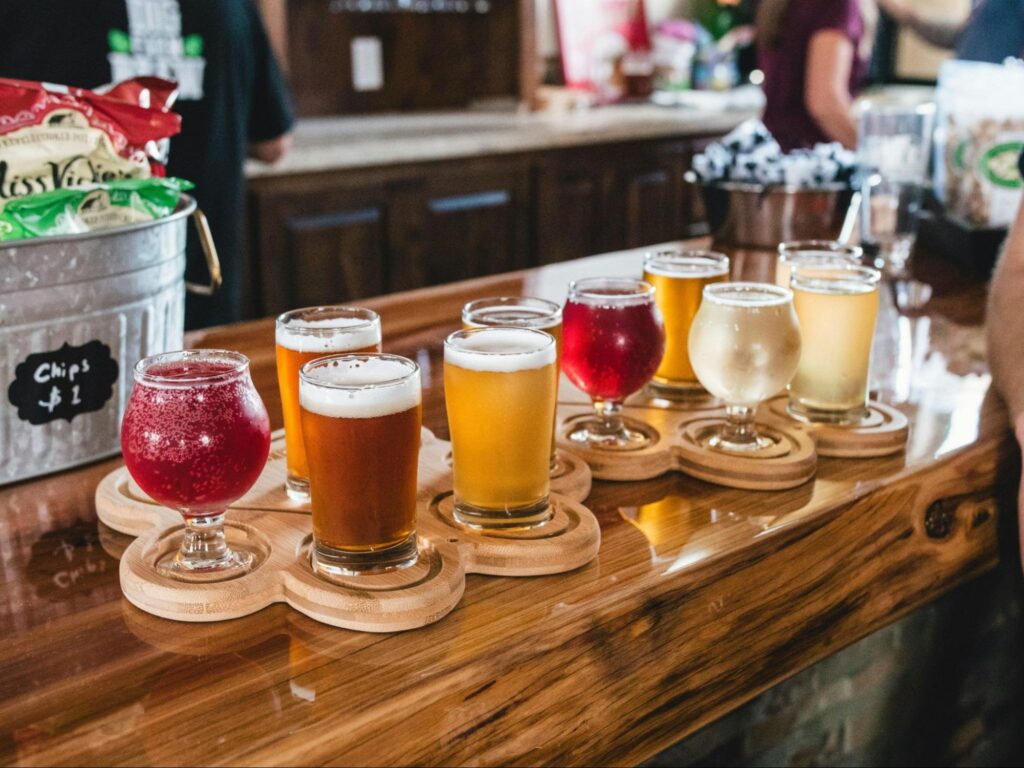 A flight of eight assorted craft beers in small glasses sits on a wooden bar counter, perfect for sampling during fun date ideas NJ, with snacks and patrons visible in the background.