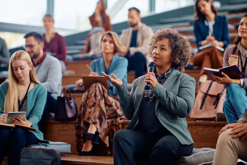 A woman holding a microphone speaks while seated among other attendees at a corporate event in an auditorium-style seminar. People around her are taking notes and listening attentively, exploring new event locations and ideas for Tac Ops NJ.