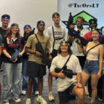 A group of twelve young adults pose indoors with laser tag gear, wearing headbands and holding laser tag guns, in front of a banner that reads "@TacOpsLT," one of the top kids birthday party places New Jersey has to offer.