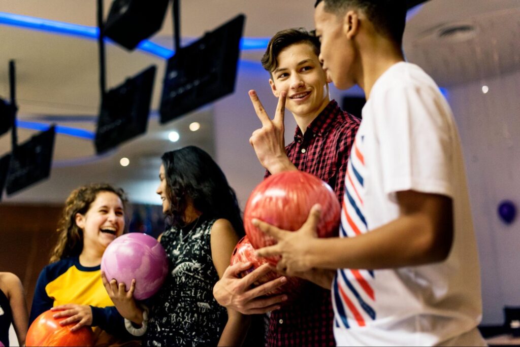 Four teenagers holding bowling balls stand together at a bowling alley, smiling and talking; one teen makes a peace sign with his fingers—a perfect spot for indoor fun and birthday party ideas in New Jersey.