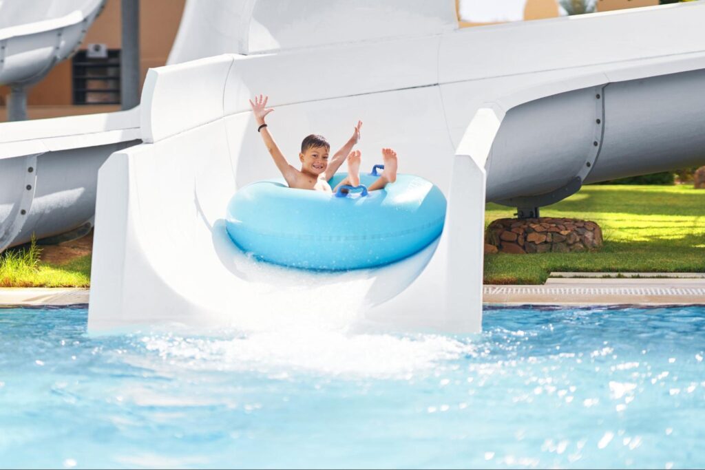 Two children on a blue inflatable raft exit a white water slide, splashing into a pool with their arms raised—perfect for indoor fun and birthday party ideas for kids in New Jersey.