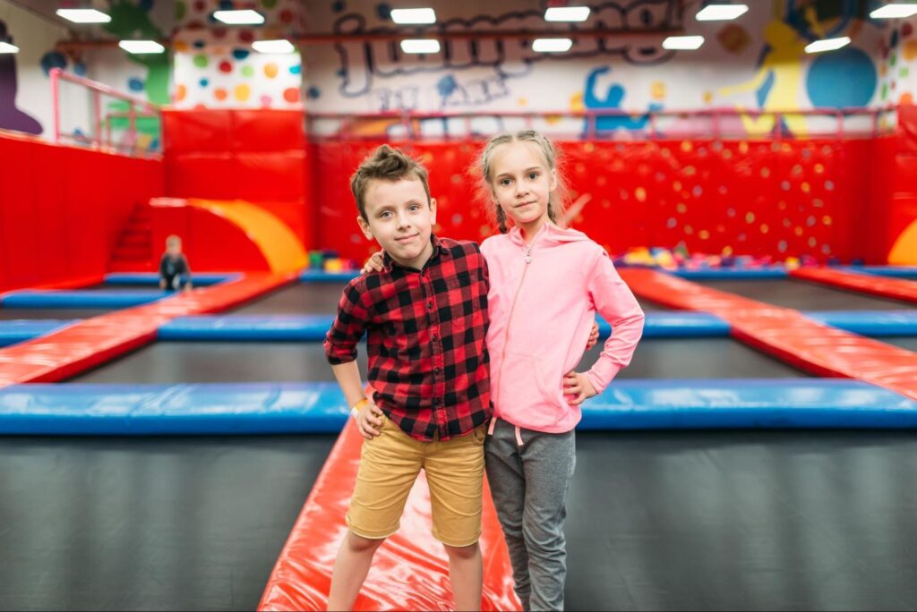 Two children stand side by side on a trampoline walkway in an indoor trampoline park with red, blue, and colorful decor—perfect for fun birthday party ideas or an unforgettable indoor party for kids in New Jersey.
