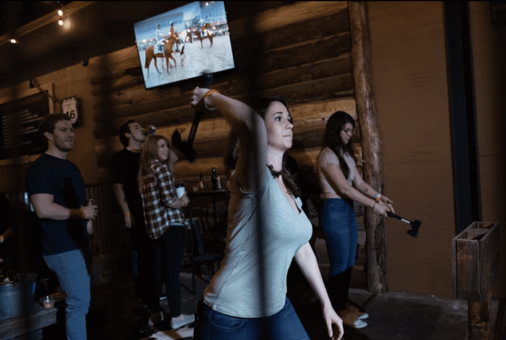 A woman throws an axe at an indoor axe-throwing venue in New Jersey while others watch and wait their turn—a fun spot for Halloween date night, complete with a TV showing a horse race mounted on the wall.