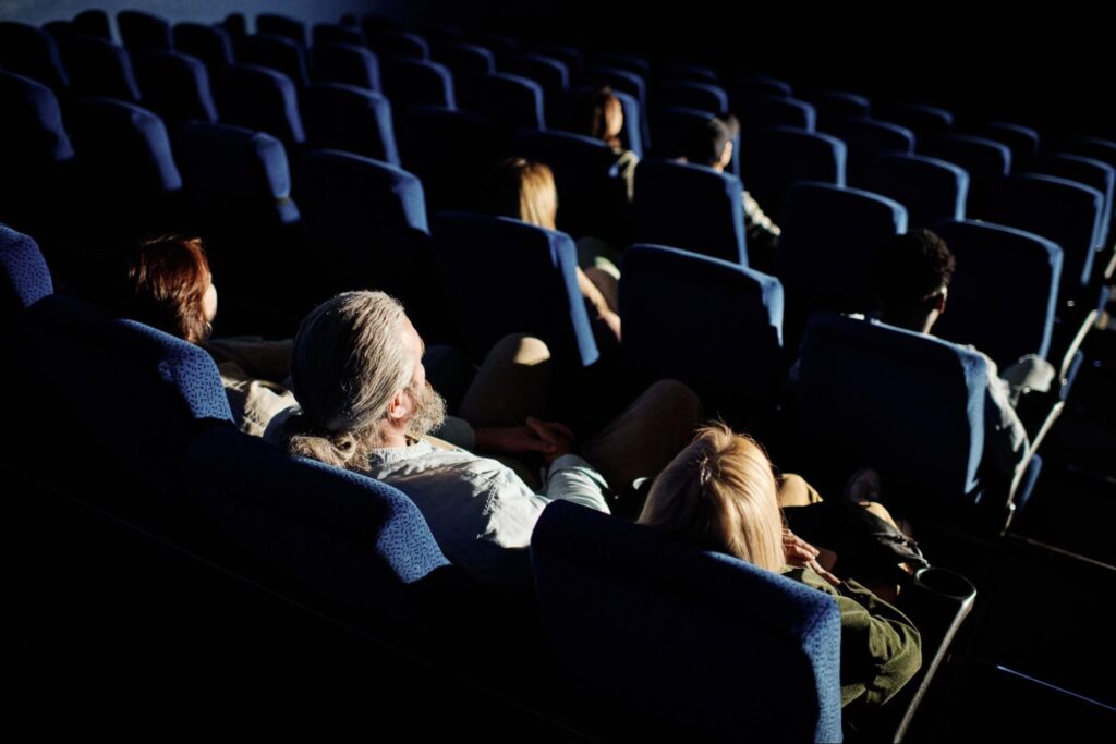 A small group of people sit spaced apart in dark blue theater seats, watching a movie in a dimly lit New Jersey cinema—perfect for a cozy Halloween date night.