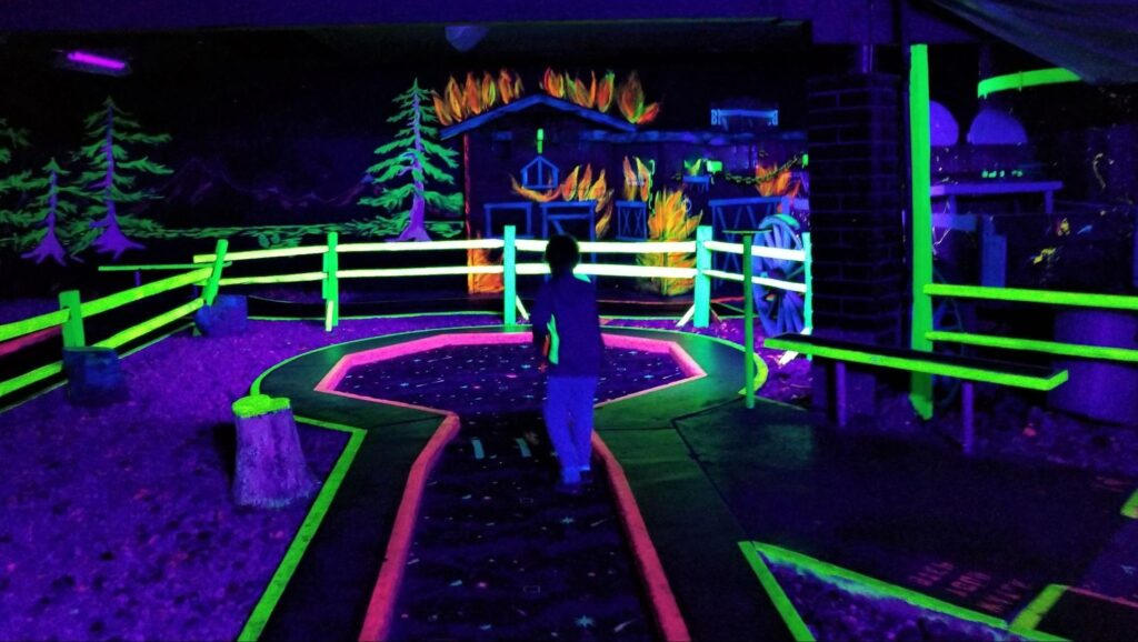 A child stands on a glow-in-the-dark mini golf course, with neon-painted obstacles and a mural of a burning house and trees in the background—one of New Jersey’s most unique winter date ideas for couples seeking fun date ideas indoors.