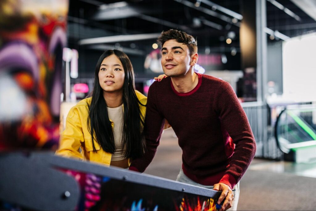 Two young adults, one in a yellow jacket and the other in a red sweater, play a pinball machine together in an arcade—one of the best fun dates or winter date ideas for couples in New Jersey.