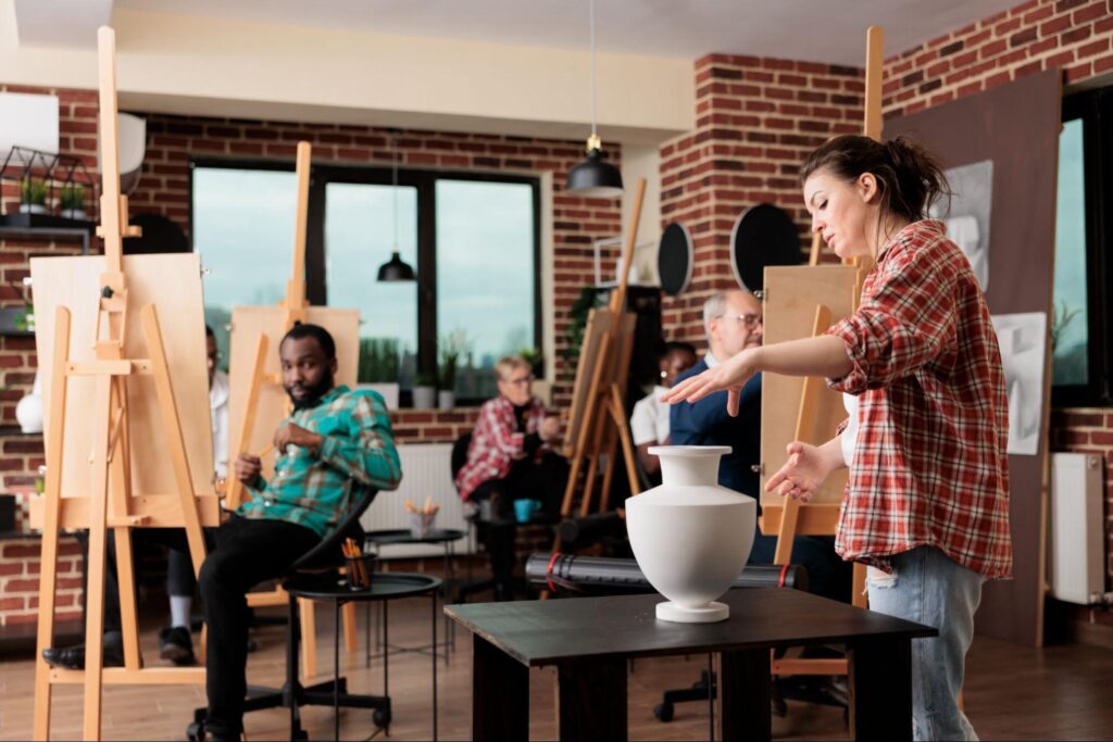 A woman stands beside a white ceramic vase on a table, while people seated at easels sketch in an art studio with brick walls and large windows—an inspiring setting for creative corporate event ideas in New Jersey.