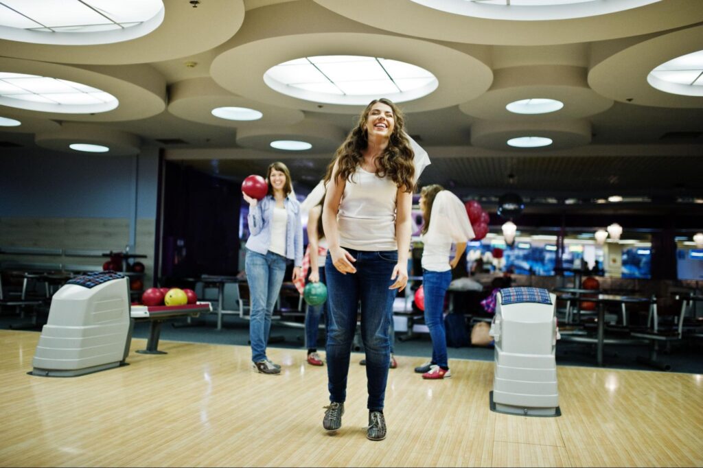 A woman smiles while standing on a bowling lane in New Jersey, with others holding bowling balls in the background—making it one of the best corporate events or team-building corporate event ideas.