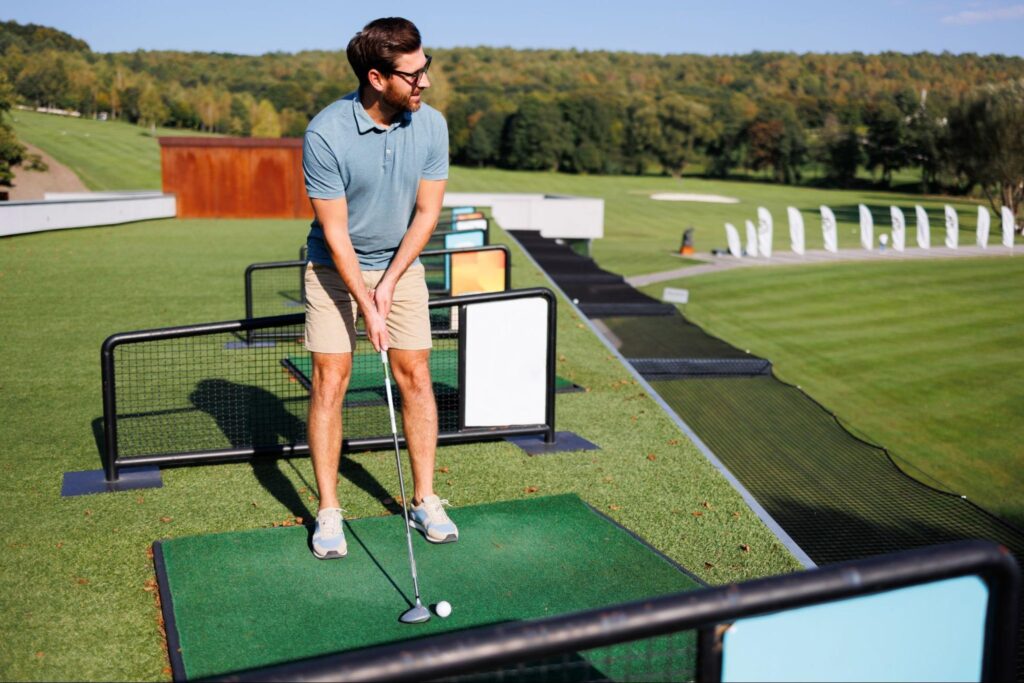 A man in a blue shirt and khaki shorts stands on a driving range mat in New Jersey, holding a golf club and preparing to hit a golf ball. The range and golf course are visible—perfect inspiration for corporate event ideas.