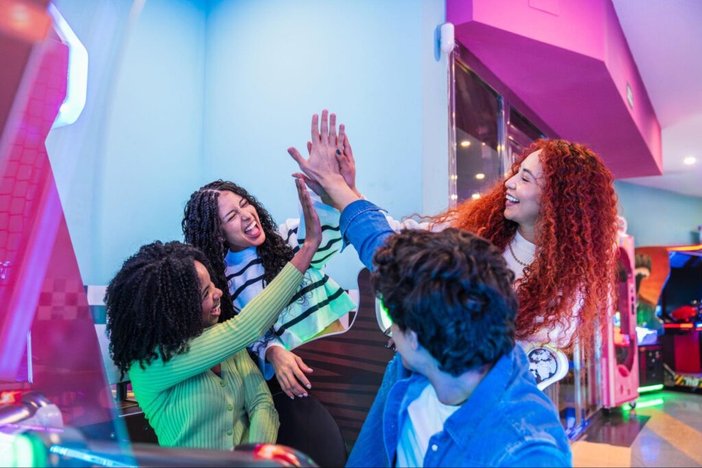 Four young adults in a brightly lit New Jersey arcade celebrate together, smiling and giving each other high-fives in front of arcade machines—perfect inspiration for fun corporate event ideas.