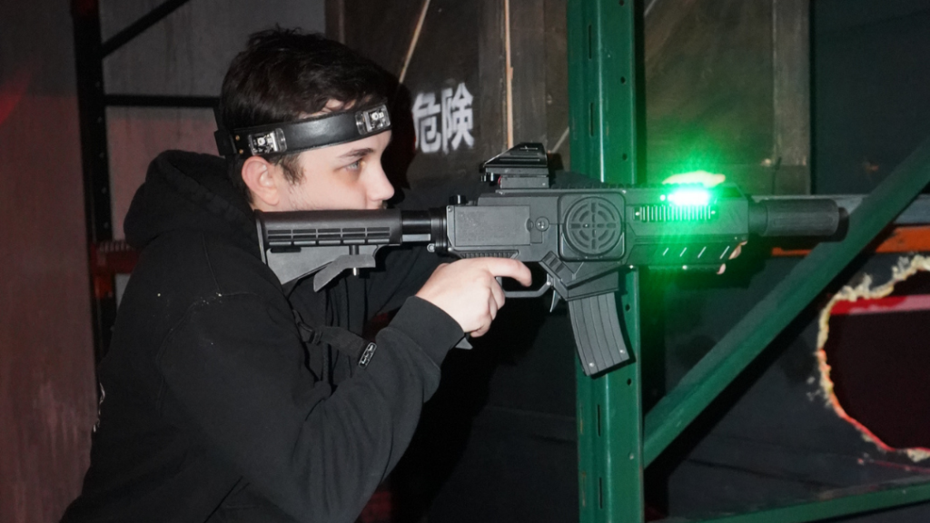 A person wearing a headband aims a toy laser gun with a green light in an indoor, dimly lit setting, capturing the excitement of tactical laser tag and the thrill of a memorable group experience.