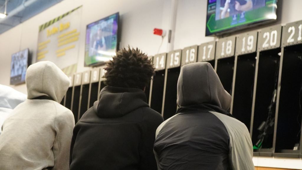 Three people in hooded jackets sit before numbered lockers, watching TV screens on the wall—capturing the anticipation of a Tactical Laser Tag group experience.