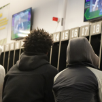 Three people in hooded jackets sit before numbered lockers, watching TV screens on the wall—capturing the anticipation of a Tactical Laser Tag group experience.