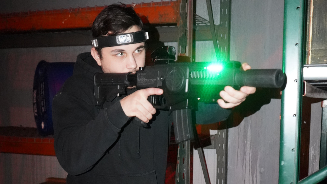 A person wearing a headband aims a laser tag gun with a green light in an indoor setting with metal shelves, perfect for corporate team building activities.