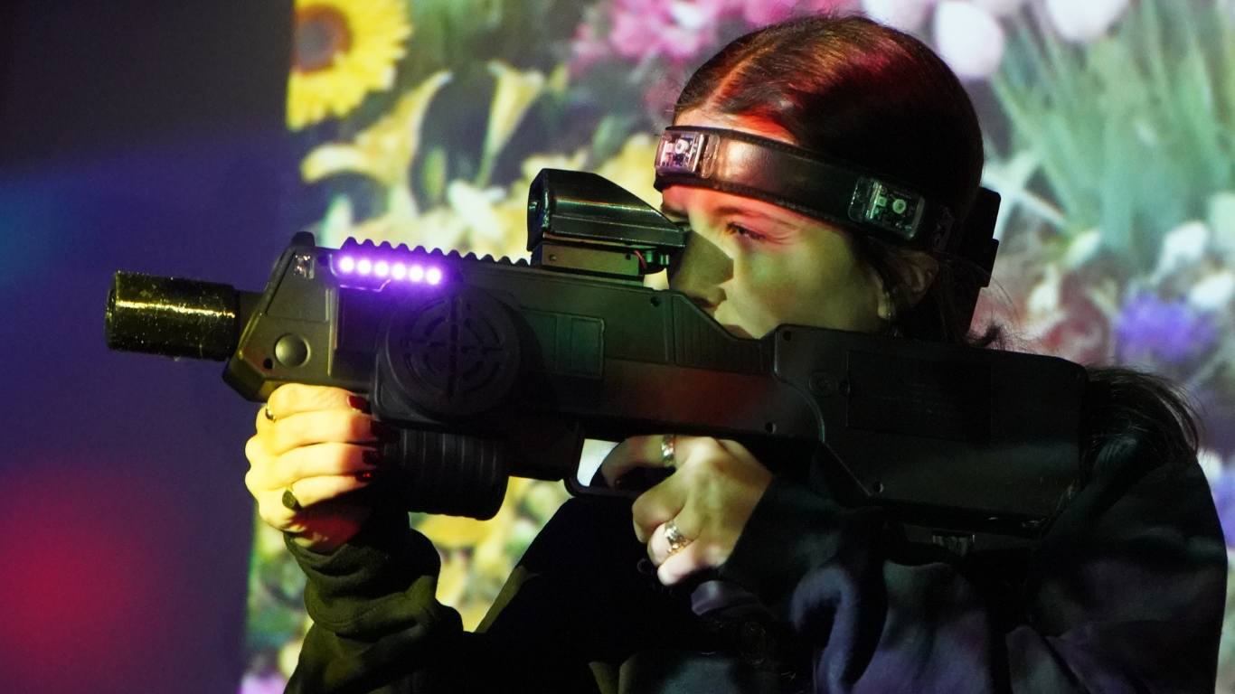 A person wearing a headband aims a black laser tag gun, with a background featuring colorful flowers—perfect for a fun corporate team building activity.