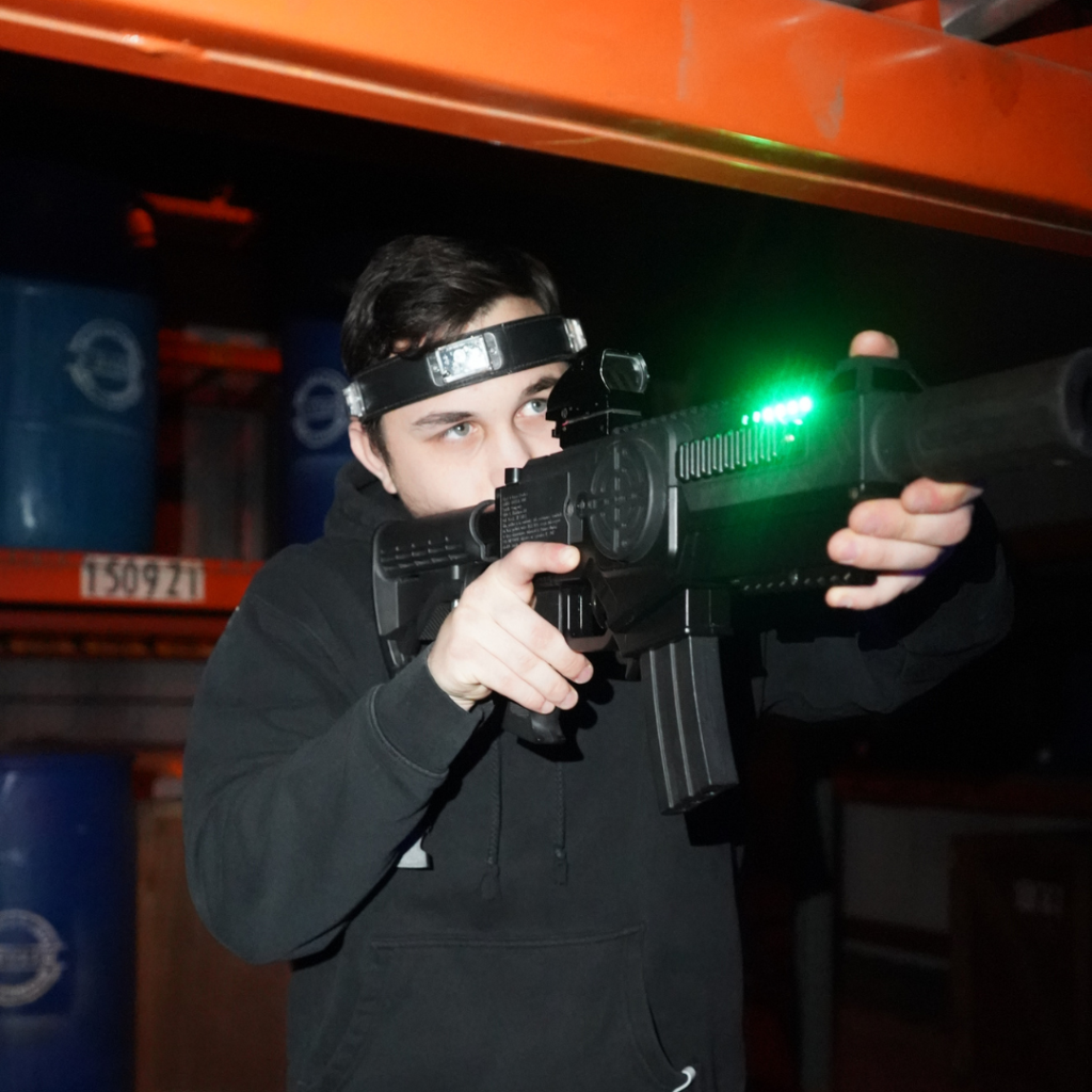 A person wearing a headband aims a laser tag gun with a green light in an indoor Laser Tag in Fairfield arena filled with metal shelves and blue barrels.