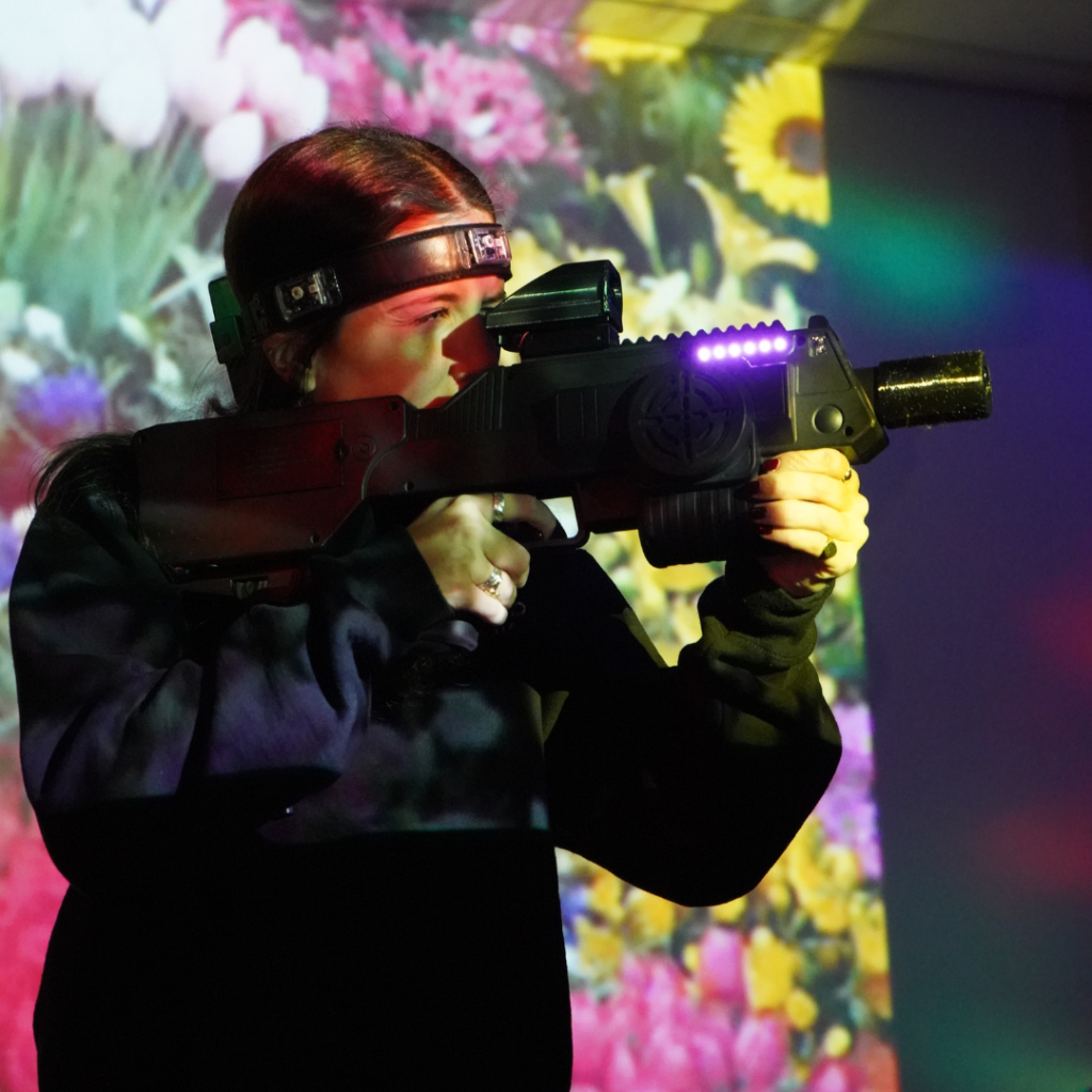 A person enjoying Laser Tag in Florham Park wears a headband and aims a laser tag gun in a room with colorful floral projections on the wall.