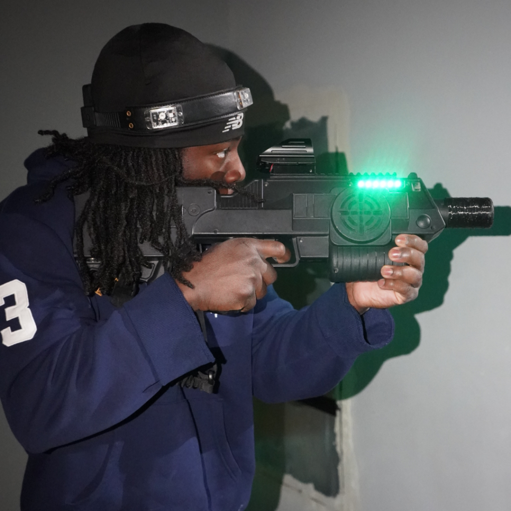 A person wearing a headband and blue hoodie aims a black laser tag gun with green lights during an exciting game of Laser Tag in Princeton.