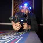 A person wearing a glowing headband holds a laser tag gun and crouches behind a table in a dimly lit room.