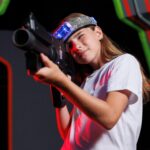 A person wearing a headband with lights aims a laser tag gun, standing in an indoor laser tag arena with colorful neon decorations during an exciting summer camp field trip.